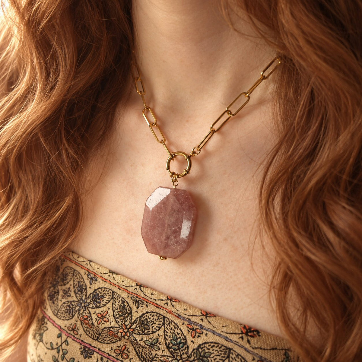A close-up of a woman wearing a gold chain necklace with a faceted pink strawberry quartz stone pendant, styled with a boho patterned top.