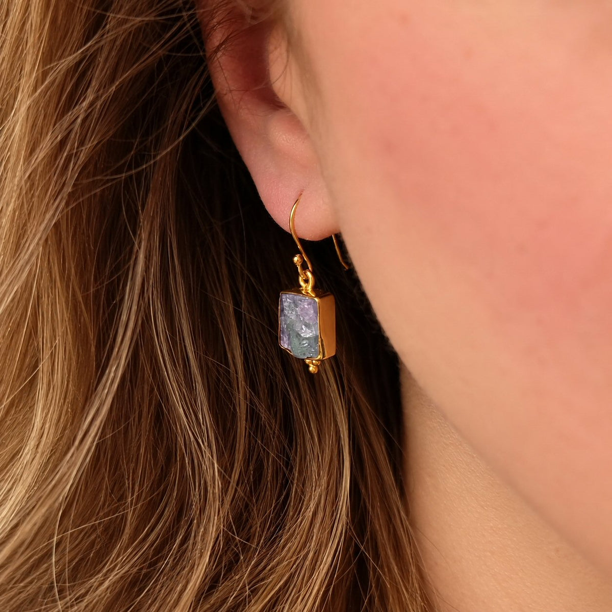 A close-up of a person's ear wearing a gold dangle earring with a rectangular iridescent stone.
