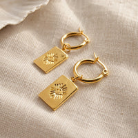 A pair of gold hoop earrings with rectangular sunburst eye charms, displayed on a textured beige linen fabric background.